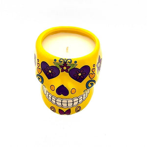 The Small Yellow Ceramic Skull Candle is a white soy wax candle in a yellow ceramic holder, shaped like a sugar skull and adorned with purple and red floral designs. It features a cozy nutmeg scent.