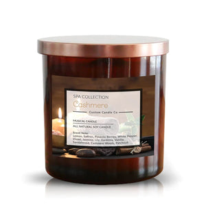 The Musical Spa Cashmere Candle, in a brown glass jar with metallic lid, offers relaxing linen, saffron, vanilla, and patchouli notes for a soothing spa experience.