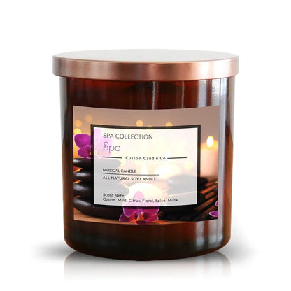 The "Musical Spa The Spa Candle" is an all-natural soy candle in a brown glass jar with a metallic lid, featuring purple orchids and spa stones—ideal for those seeking natural scented candles with relaxing fragrance notes.