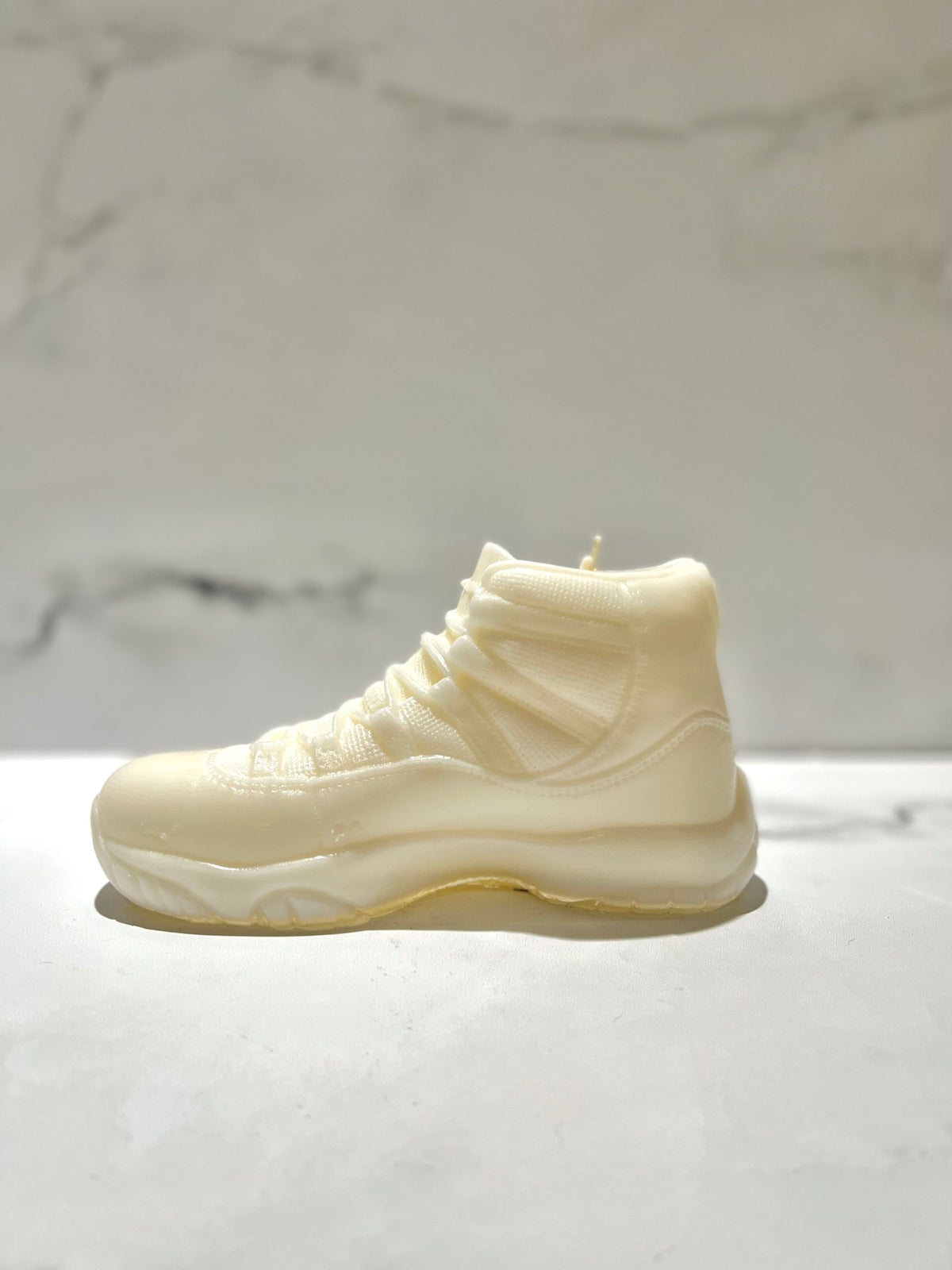 The AJ-11 White Hightop Sneaker Candle – Large (On Backorder), hand poured and infused with Black Ice fragrance, rests on a white surface against a light marble background.
