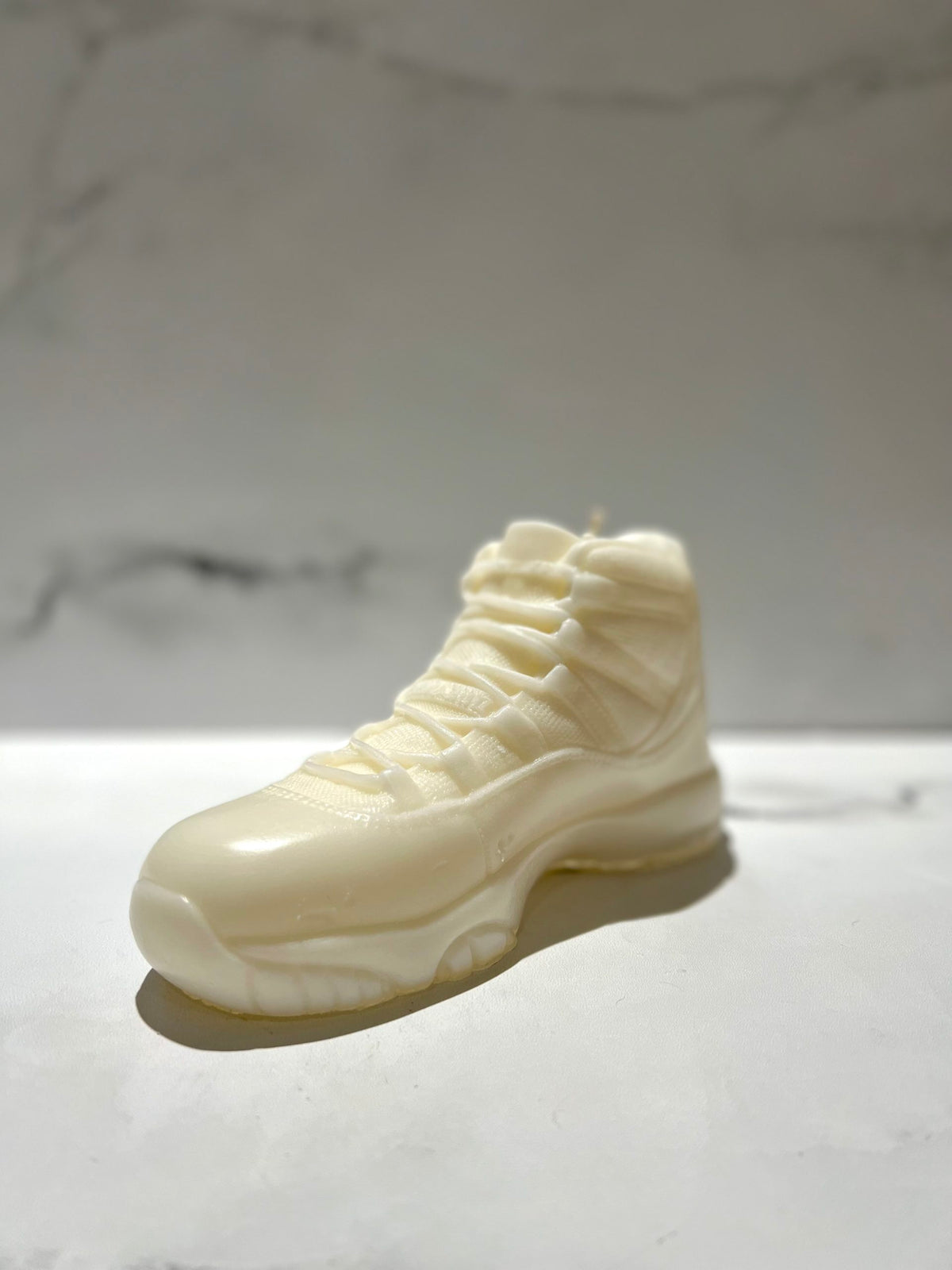 The AJ-11 White Hightop Sneaker Candle – Large (On Backorder) is shown on a light surface against a marble-patterned background.