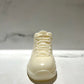 The AJ-11 White Hightop Sneaker Candle – Large (On Backorder) stands upright on a marble surface, showcasing its hand-poured white design against a blurred background.