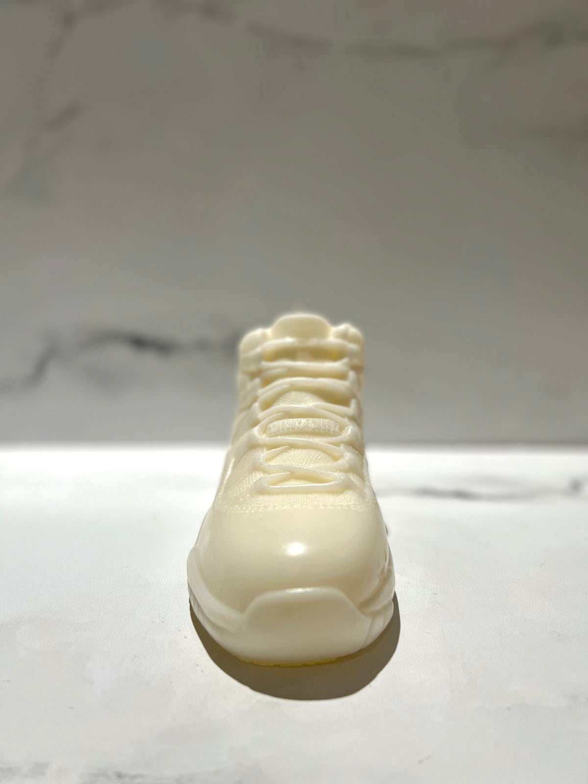 The AJ-11 White Hightop Sneaker Candle – Large (On Backorder) stands upright on a marble surface, showcasing its hand-poured white design against a blurred background.