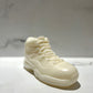 The AJ-11 White Hightop Sneaker Candle – Large (On Backorder) is a hand-poured candle shaped like a classic white high-top sneaker, displayed on a smooth, light surface with a softly blurred marble-patterned background.