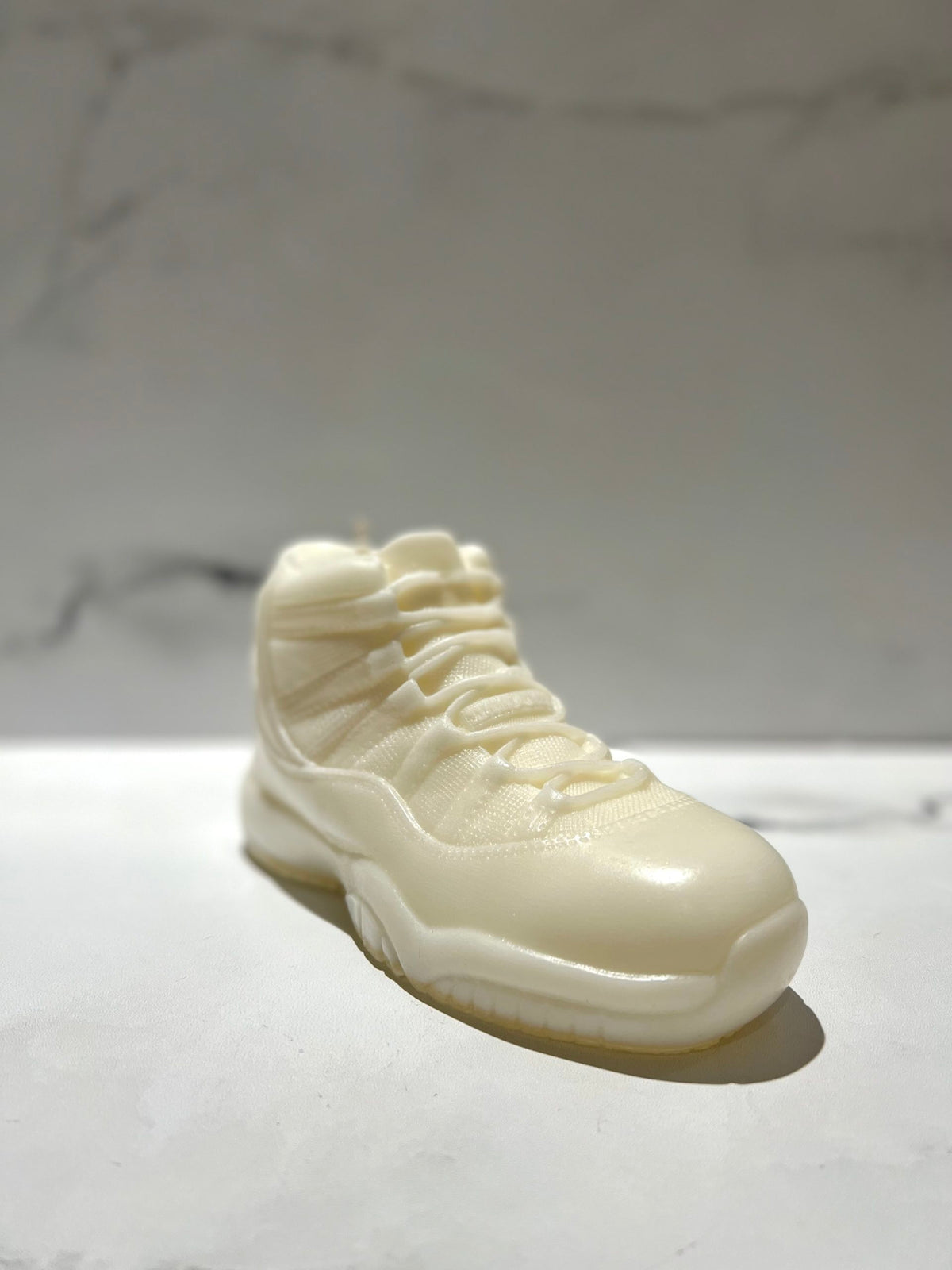 The AJ-11 White Hightop Sneaker Candle – Large (On Backorder) is a hand-poured candle shaped like a classic white high-top sneaker, displayed on a smooth, light surface with a softly blurred marble-patterned background.