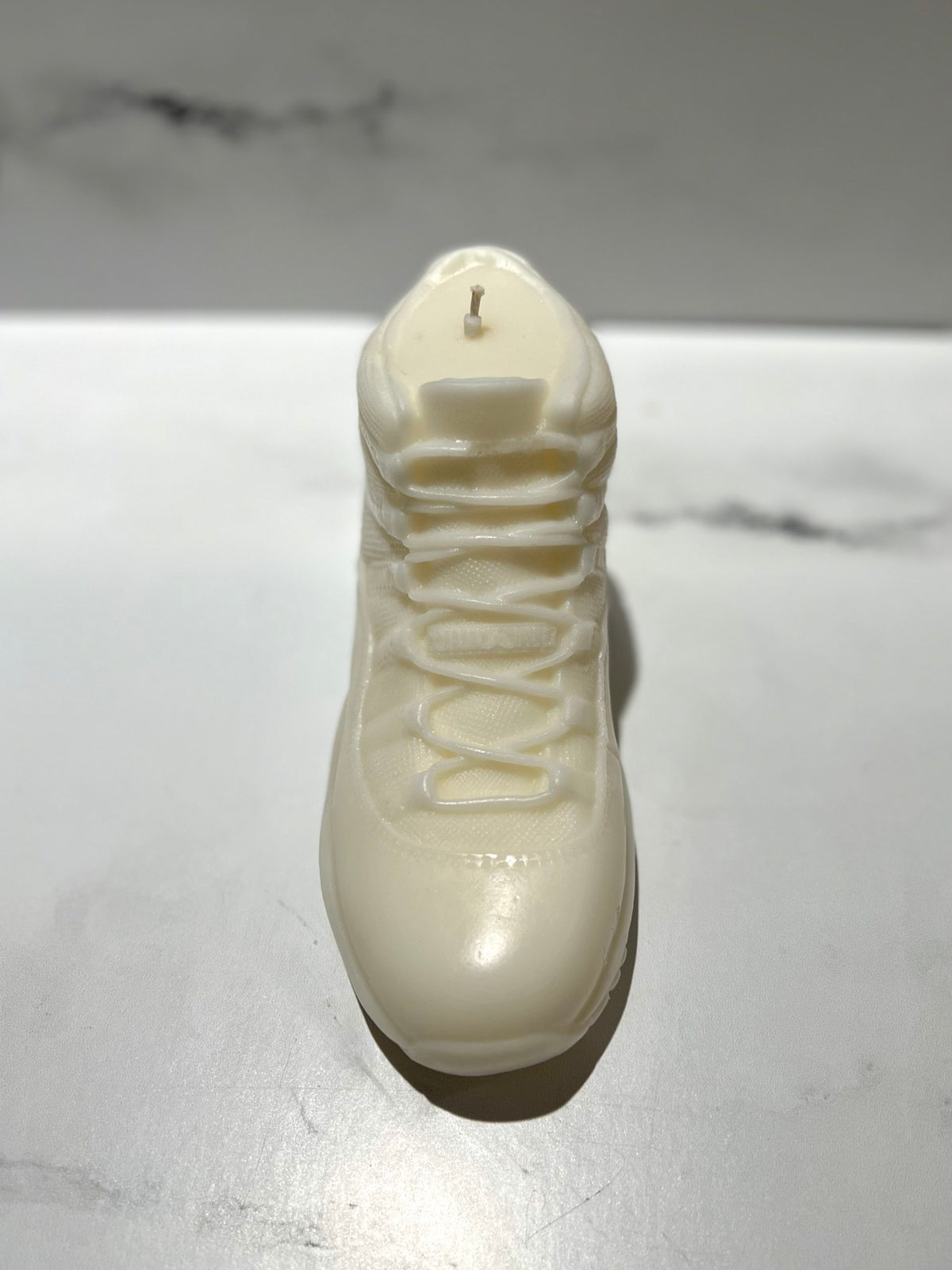 The AJ-11 White Hightop Sneaker Candle – Large (On Backorder), hand poured with a Black Ice scent, rests on a smooth light surface against a marble backdrop.