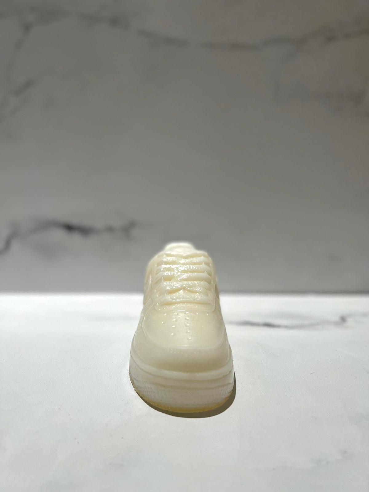 The AF1 – White Lowtop Sneaker Candle – Large (On Backorder) features a sneaker-shaped white candle on a marble surface, set against a marble-patterned background, and offers the refreshing scent of Black Ice fragrance.