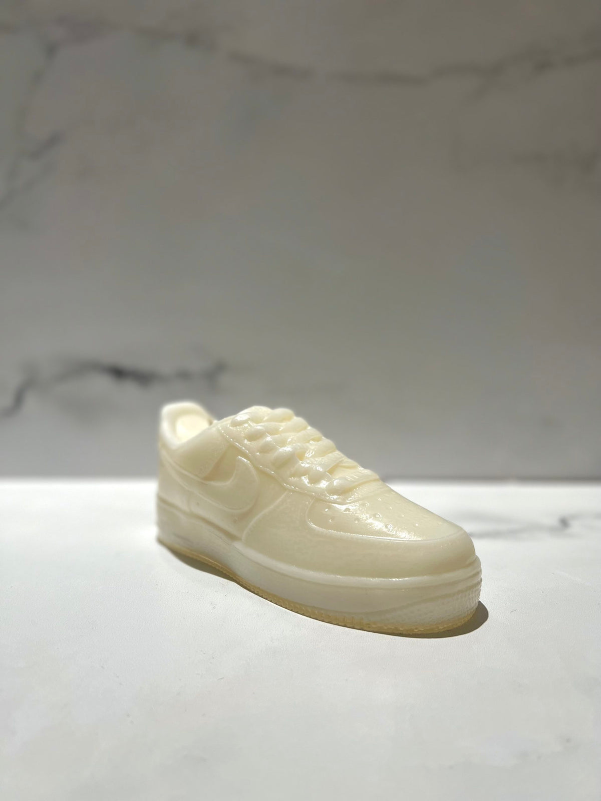 The AF1 – White Lowtop Sneaker Candle – Large (On Backorder), designed to resemble a classic white lowtop sneaker, rests on a light marble surface with a blurred marble-patterned background.