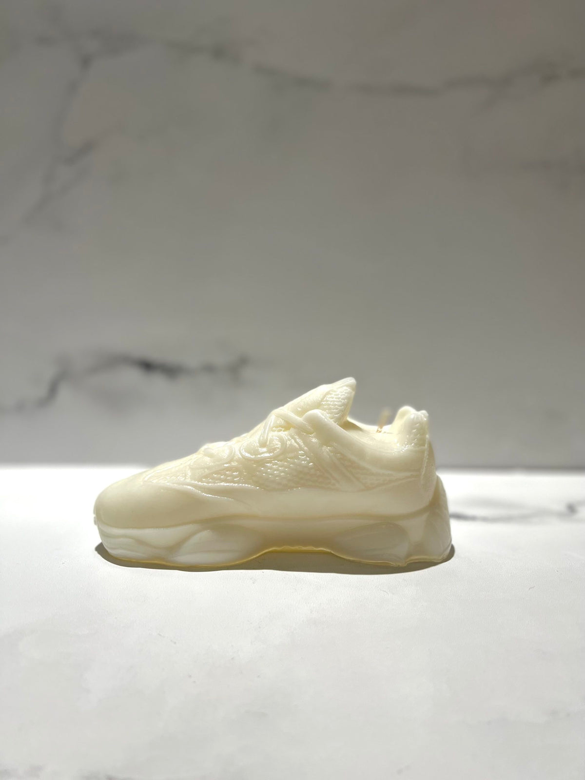 The Y500 – White Sneaker Candle – Large (On Backorder) is shown on a light marble surface with a blurred marble-patterned background, highlighting the distinct appeal of this hand-poured candle.