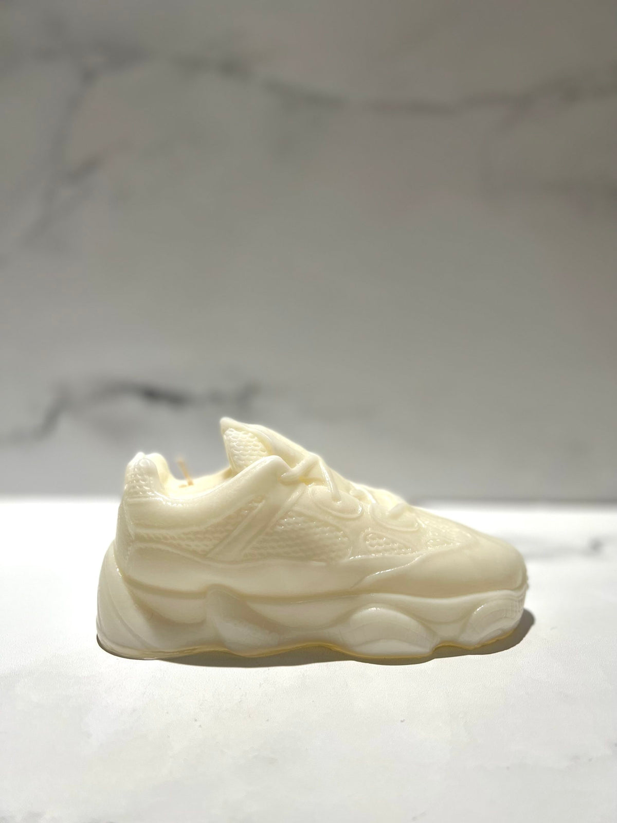 The Y500 – White Sneaker Candle – Small (On Backorder) sits on a white surface with a blurred light background, hand poured in an off-white shade and infused with Black Ice fragrance.