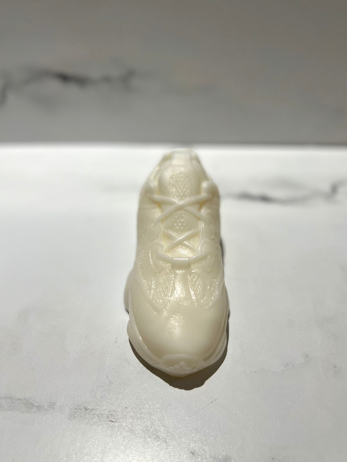 The Y500 – White Sneaker Candle – Small (On Backorder), a hand-poured, white sneaker-shaped candle, is displayed on a marble-textured surface.