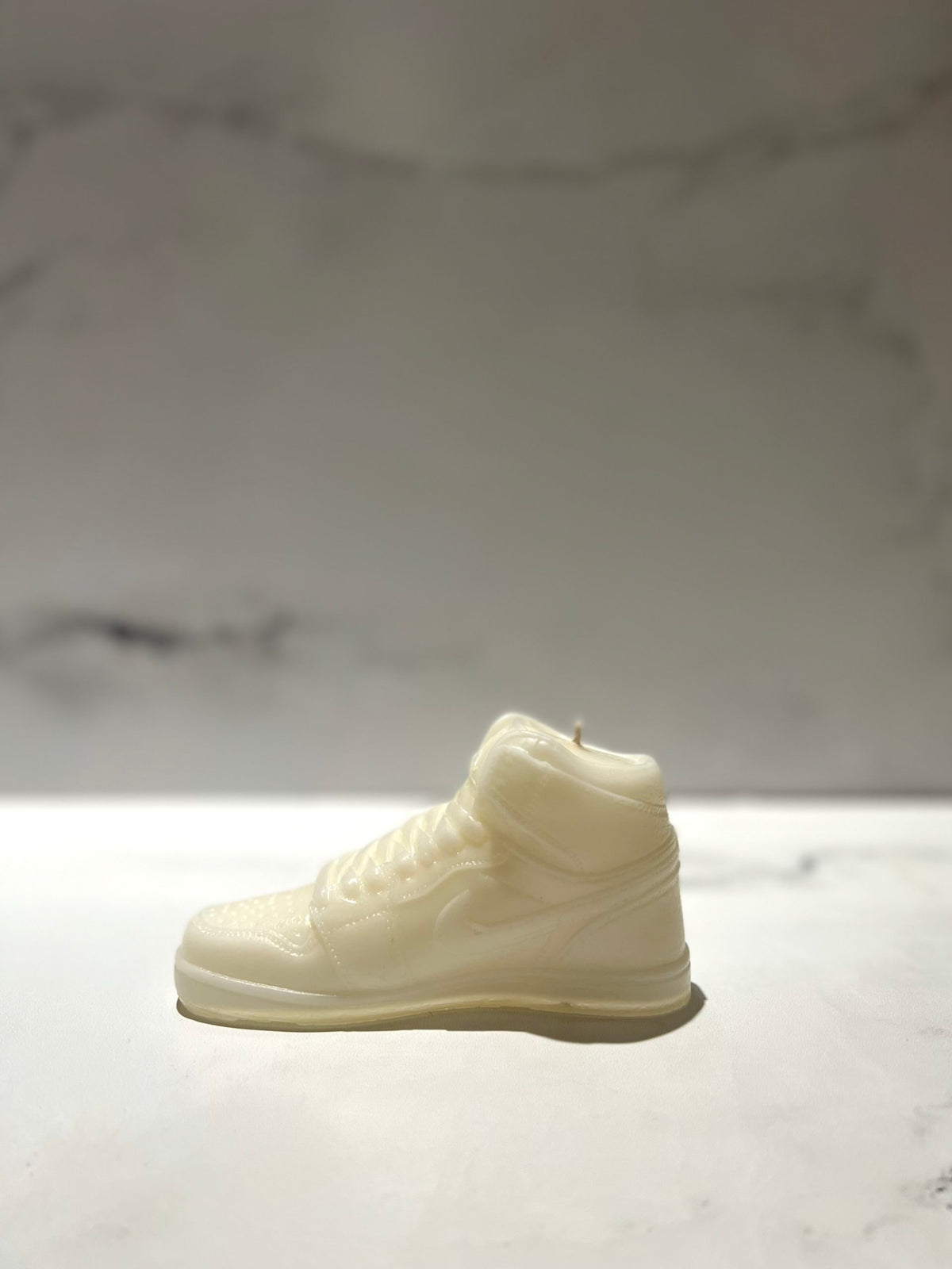 The AJ-1 White Hightop Sneaker Candle – Small (on backorder), hand poured from all natural wax, is displayed on a light surface with a blurred marble background.