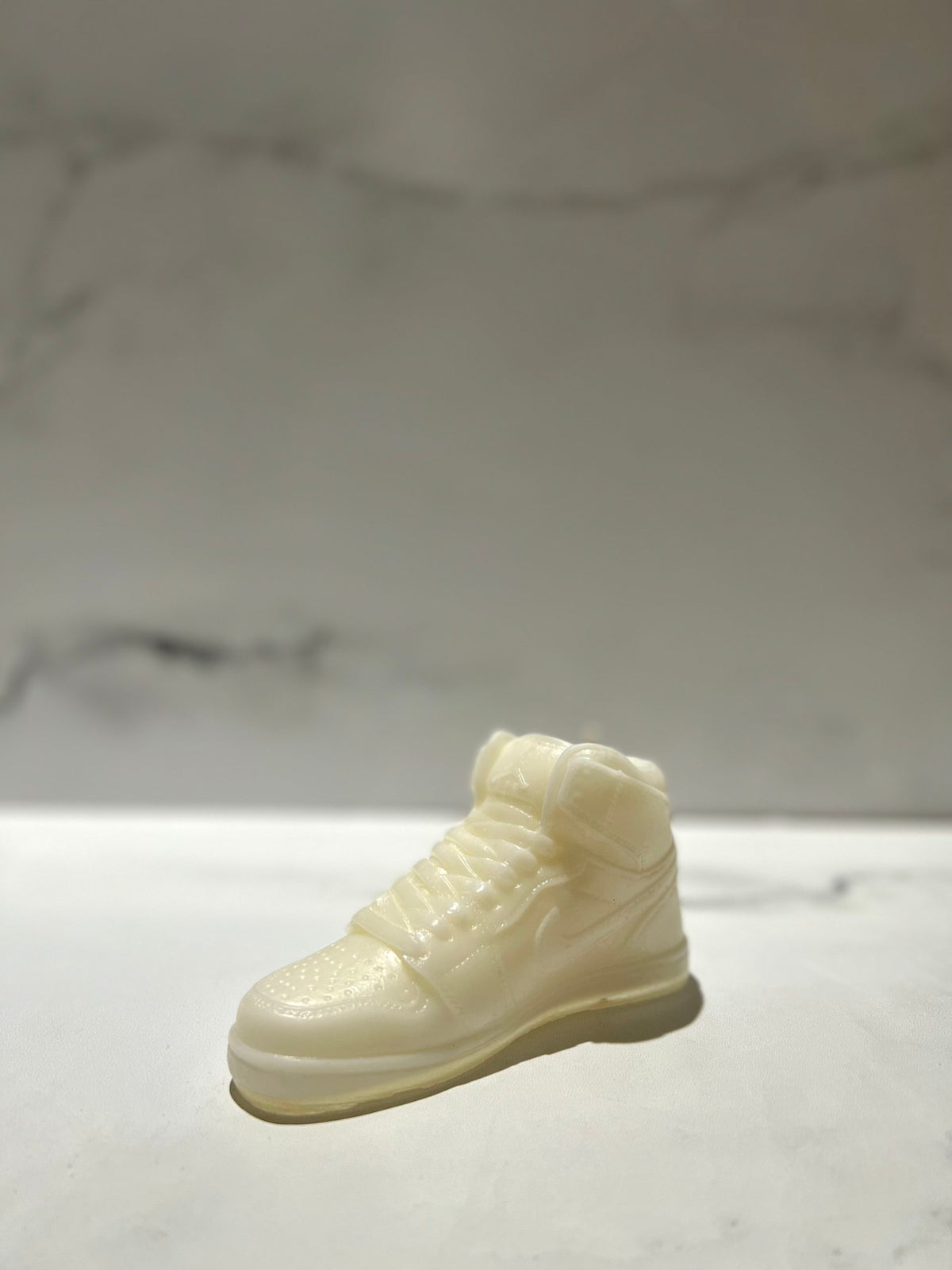 The AJ-1 White Hightop Sneaker Candle – Small (on backorder) is a realistic sneaker candle, hand-poured from all natural wax, displayed on a smooth, light surface with a marbled background.