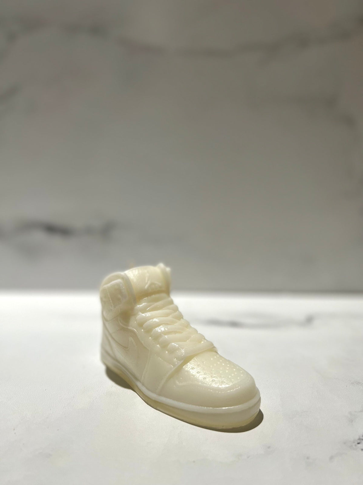 The AJ-1 White Hightop Sneaker Candle – Large (On Backorder) is a hand-poured candle shaped like a high-top sneaker, crafted from a smooth white material and displayed on a light surface with a blurred background.