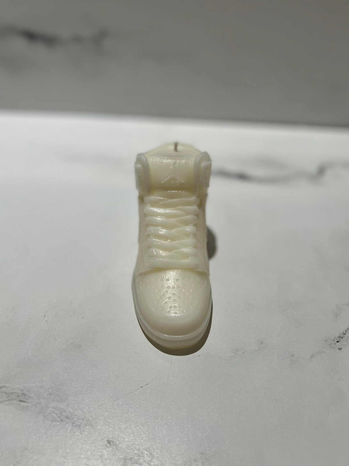 The AJ-1 White Hightop Sneaker Candle – Large (currently on backorder) is a glossy, hand-poured white candle shaped like a sneaker, set on a white marble surface and subtly scented with Black Ice fragrance.