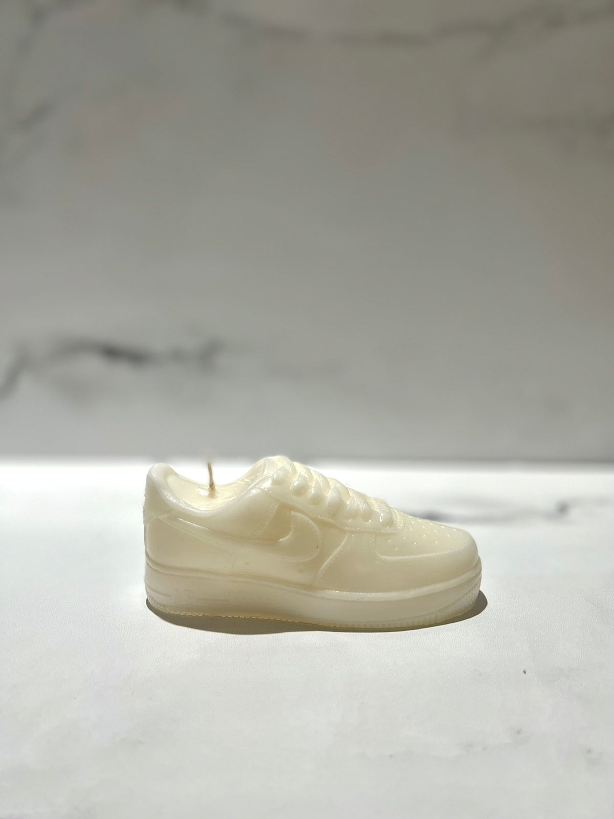 The AF1 – White Lowtop Sneaker Candle – Small (On Backorder) features a fresh Black Ice scent and a unique white design, showcased on a marble surface with its details standing out against the soft, blurred background.