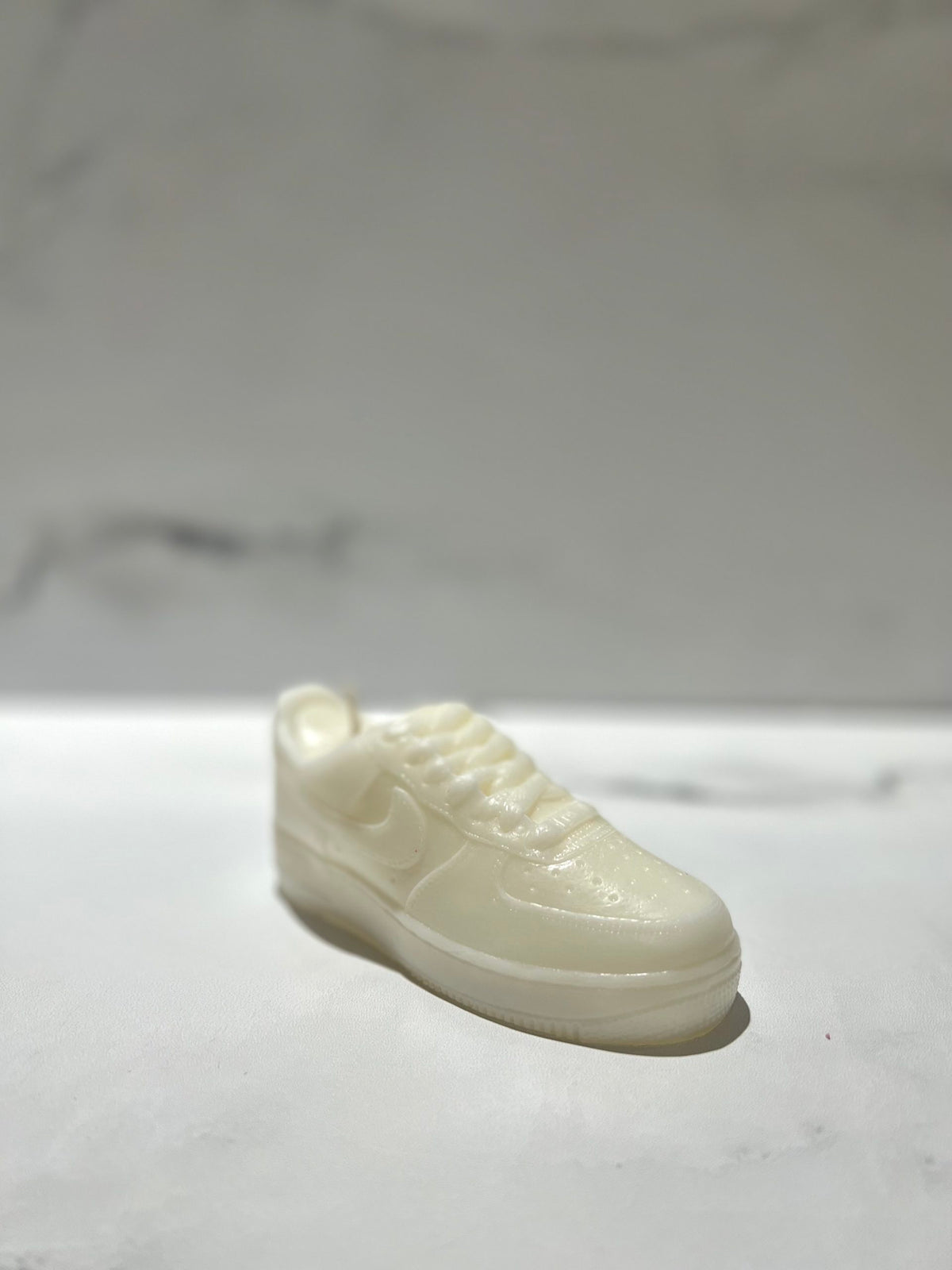 The AF1 – White Lowtop Sneaker Candle – Small (On Backorder) is a hand-poured candle shaped like a classic sneaker, featuring the fresh scent of Black Ice. It is displayed on a white surface with a blurred background.