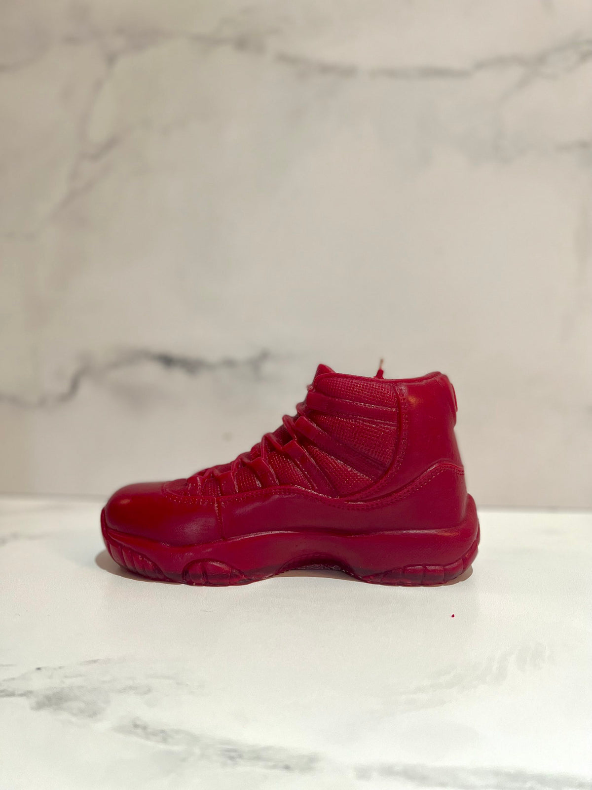 The AJ-11 Red Hightop Sneaker Candle – Large (On Backorder) is a hand-poured, all-natural wax candle shaped like a red high-top sneaker, displayed on a white surface with a light marbled background.