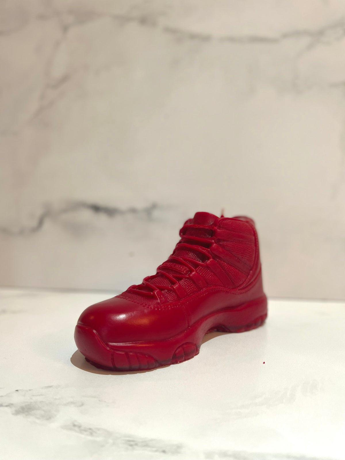 The AJ-11 Red Hightop Sneaker Candle – Large (On Backorder), crafted with all-natural wax, is displayed on a white and gray marbled surface with a light marbled backdrop.