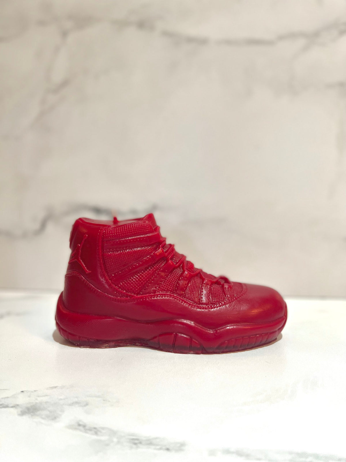 The AJ-11 Red Hightop Sneaker Candle – Large (On Backorder), a hand-poured, all-natural wax candle in a red high-top sneaker design, is shown on a white marble surface with a light marble background.