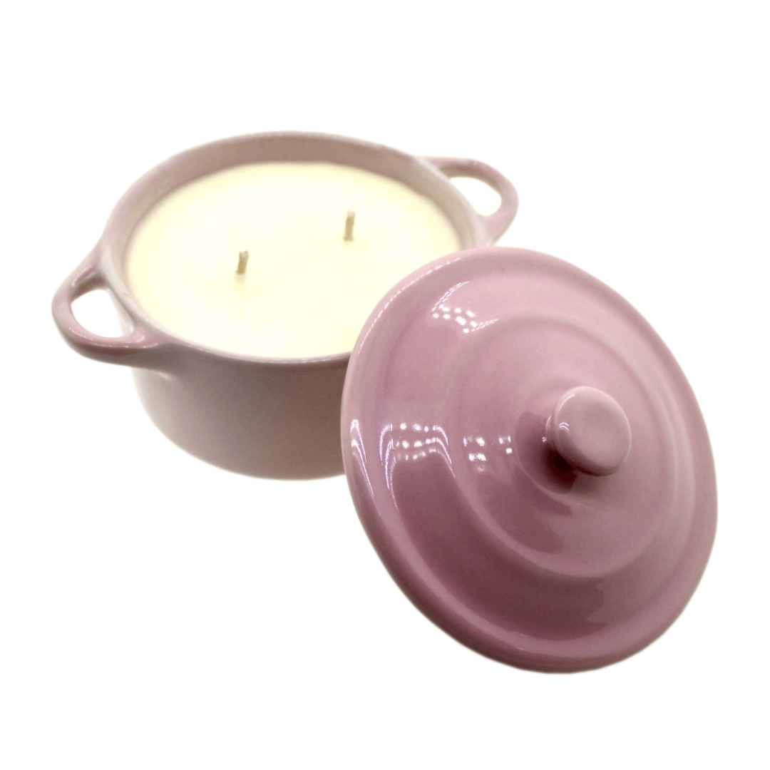 A purple ceramic Dutch oven candle with two handles, filled with white soy wax and two wicks, scented like banana nut bread, with its matching lid placed beside it.