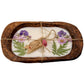 A rectangular wooden tray holds the Purple Floral Wood Candle – Floral Scented, a coconut-apricot wax tablet with pressed purple flowers and green leaves, tied with twine and a tag for a charming floral accent.