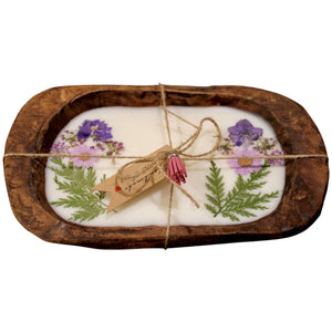 A rectangular wooden tray holds the Purple Floral Wood Candle – Floral Scented, a coconut-apricot wax tablet with pressed purple flowers and green leaves, tied with twine and a tag for a charming floral accent.