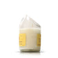 The Recycled Cognac Candle features a white coconut-apricot wax in a glass made from recycled cognac bottles, showcasing a tilted, uneven rim and yellow labels on white.