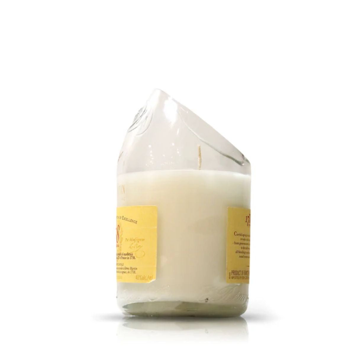 The Recycled Cognac Candle features a white coconut-apricot wax in a glass made from recycled cognac bottles, showcasing a tilted, uneven rim and yellow labels on white.