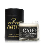 A white Recycled Azul Agave Tequila Candle in a clear glass holder labeled "Cabo Wabo Tequila Blanco" sits beside a black cylindrical Custom Candle Co. box, crafted from recycled candle materials.