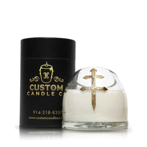The Recycled Cross of Lorraine Cognac Candle features white coconut-apricot wax in a glass holder with a gold cross, shown beside a black cylindrical Custom Candle Co. container.