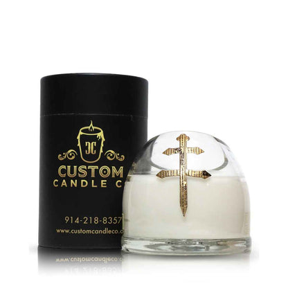 The Recycled Cross of Lorraine Cognac Candle features white coconut-apricot wax in a glass holder with a gold cross, shown beside a black cylindrical Custom Candle Co. container.