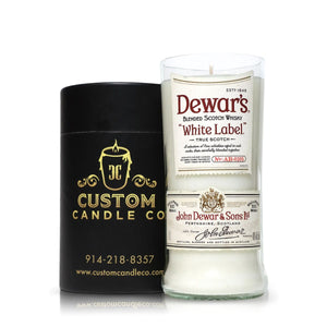 The Recycled White Label Scotch Whiskey Candle, a white candle set in an upcycled whiskey bottle glass, sits next to a black cylindrical Custom Candle Co. container.