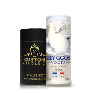 A Recycled French Vodka Candle - White, crafted from a Grey Goose bottle featuring a white goose design, sits beside a black cylindrical Custom Candle Co. container with gold text, highlighting its elegant recycled bottle craftsmanship.