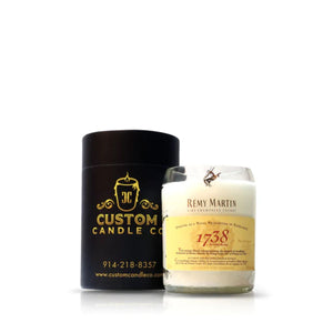 A Recycled Cognac Candle labeled "Rémy Martin 1738," made from recycled cognac bottles, sits beside a black "Custom Candle Co." cylindrical container with gold text.