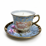 The Floral Teacup Candles feature a gold-rimmed teacup on a matching saucer, with a softly glowing soy candle beside a purple flower—ideal for adding vintage charm to your décor.