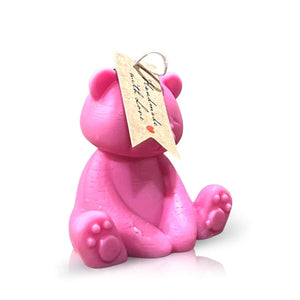 The Teddy Bear Pastel Pink Baby Candle features a tag on its head and sits on a white reflective surface.