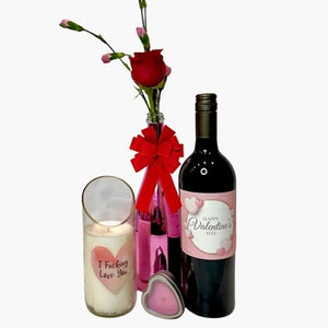 The Valentines Luxourious Gift Set – Pink/Clear Shiney set includes a pink vase with a red rose, a "Happy Valentine's Day" wine bottle, a champagne-scented "I Fucking Love You" candle, and a heart-shaped tin on a white background.