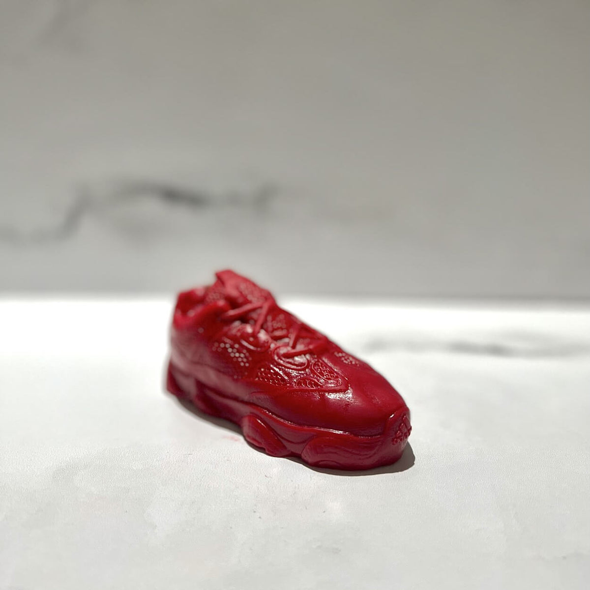 The Y500 – Red Sneaker Candle – Small (On Backorder), hand poured from natural wax, is displayed on a white, lightly textured surface against a light gray background.