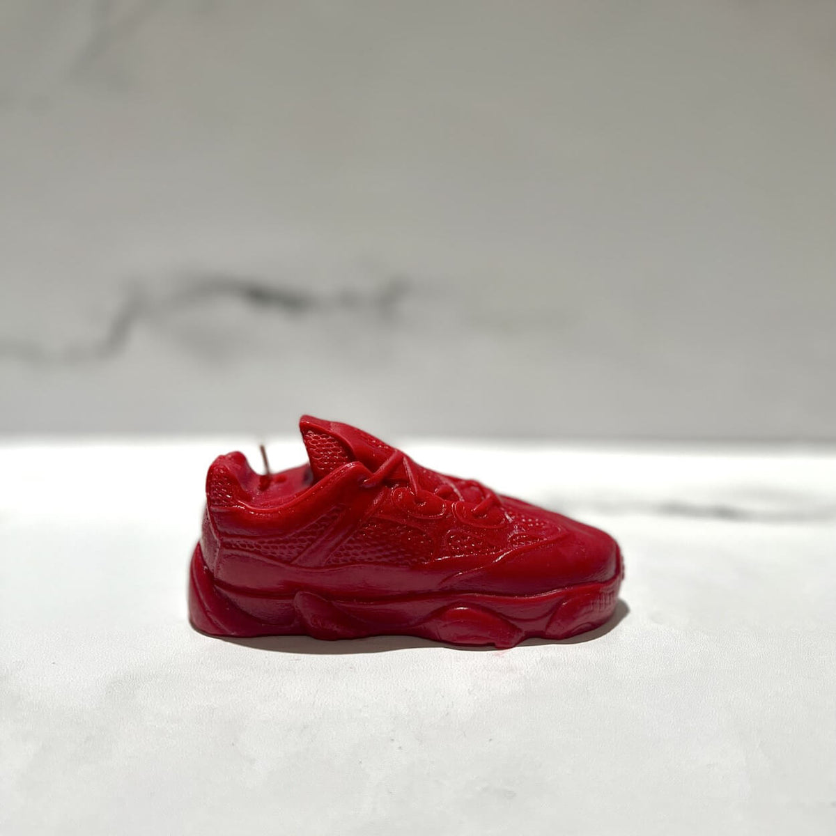 The Y500 – Red Sneaker Candle – Small (On Backorder), hand poured from natural wax, is displayed on a white surface against a blurred marble-patterned background.