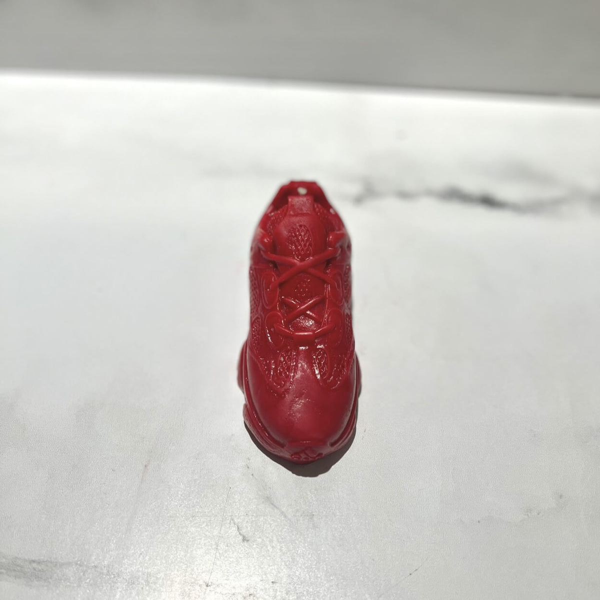 A Y500 – Red Sneaker Candle – Small (On Backorder) is placed on a white surface, casting a shadow to one side.
