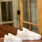 Two Y500 – White Sneaker Candle – Large (On Backorder) rest on a wooden surface, shaped like white sneakers, with a glass-paneled door in the background.