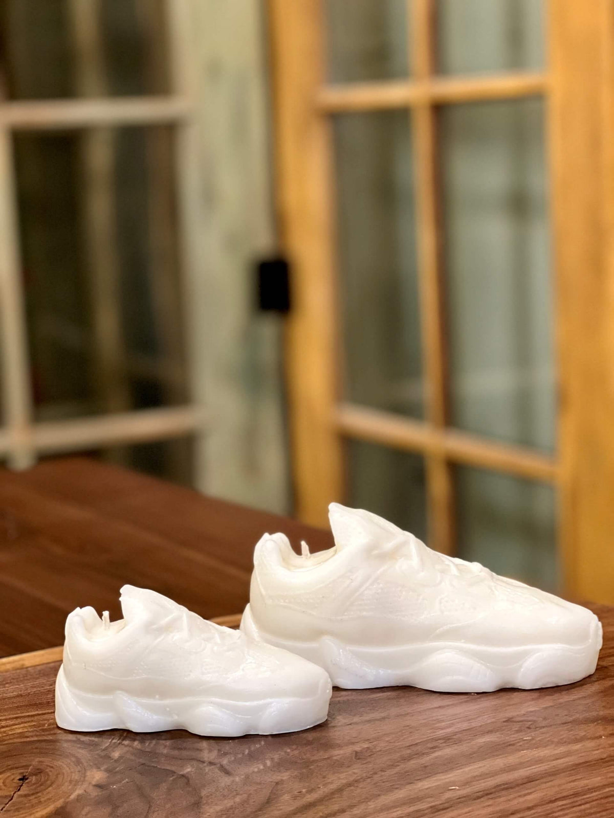 Two Y500 – White Sneaker Candle – Large (On Backorder) rest on a wooden surface, shaped like white sneakers, with a glass-paneled door in the background.