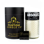 A Recycled Black Label Vodka Candle from Custom Candle Co. sits next to its sleek black and gold cylindrical packaging and a branded matchbox, all crafted from a repurposed Absolut vodka bottle.