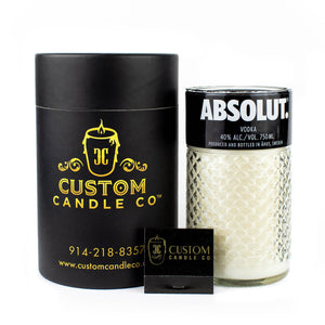 A Recycled Black Label Vodka Candle from Custom Candle Co. sits next to its sleek black and gold cylindrical packaging and a branded matchbox, all crafted from a repurposed Absolut vodka bottle.