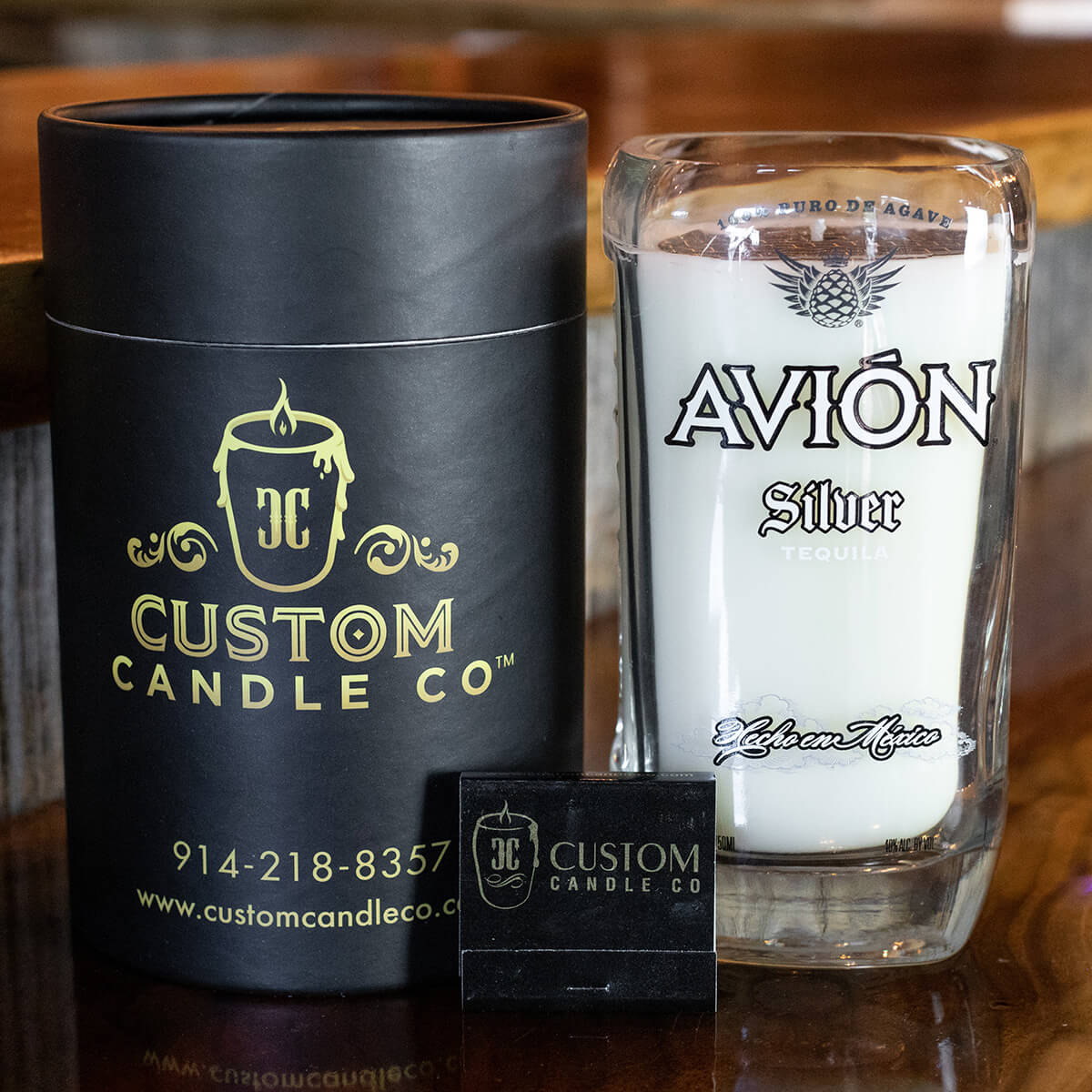 The Recycled Mexican-Style Silver Tequila Candle is displayed in a repurposed Avión Silver Tequila bottle glass, alongside sleek black cylindrical packaging and a business card—highlighting its unique recycled style.