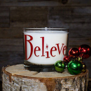 The Believe 2 Wick Candle in a glass holder with “Believe” in red letters sits on a tree stump beside red and green ornaments, bringing festive holiday scents and cheer to your decor.