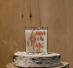 The Believe in the Magic Candle is a white natural soy candle in a glass holder with red "Believe in the Magic" text, displayed on a wooden slab with clear gems for festive charm.