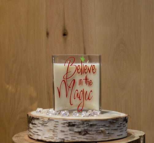 The Believe in the Magic Candle is a white natural soy candle in a glass holder with red "Believe in the Magic" text, displayed on a wooden slab with clear gems for festive charm.