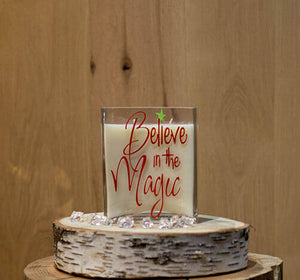 The Believe in the Magic Candle is a white natural soy candle in a glass holder with red "Believe in the Magic" text, displayed on a wooden slab with clear gems for festive charm.